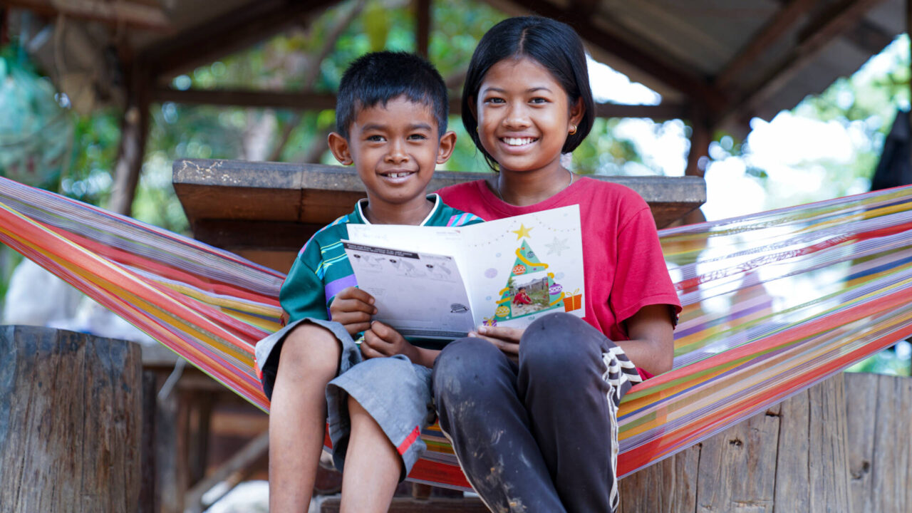 Seated closely together in a colorful, striped hammock, a young Cambodian boy and girl smile at the camera while holding a Christmas card between them.