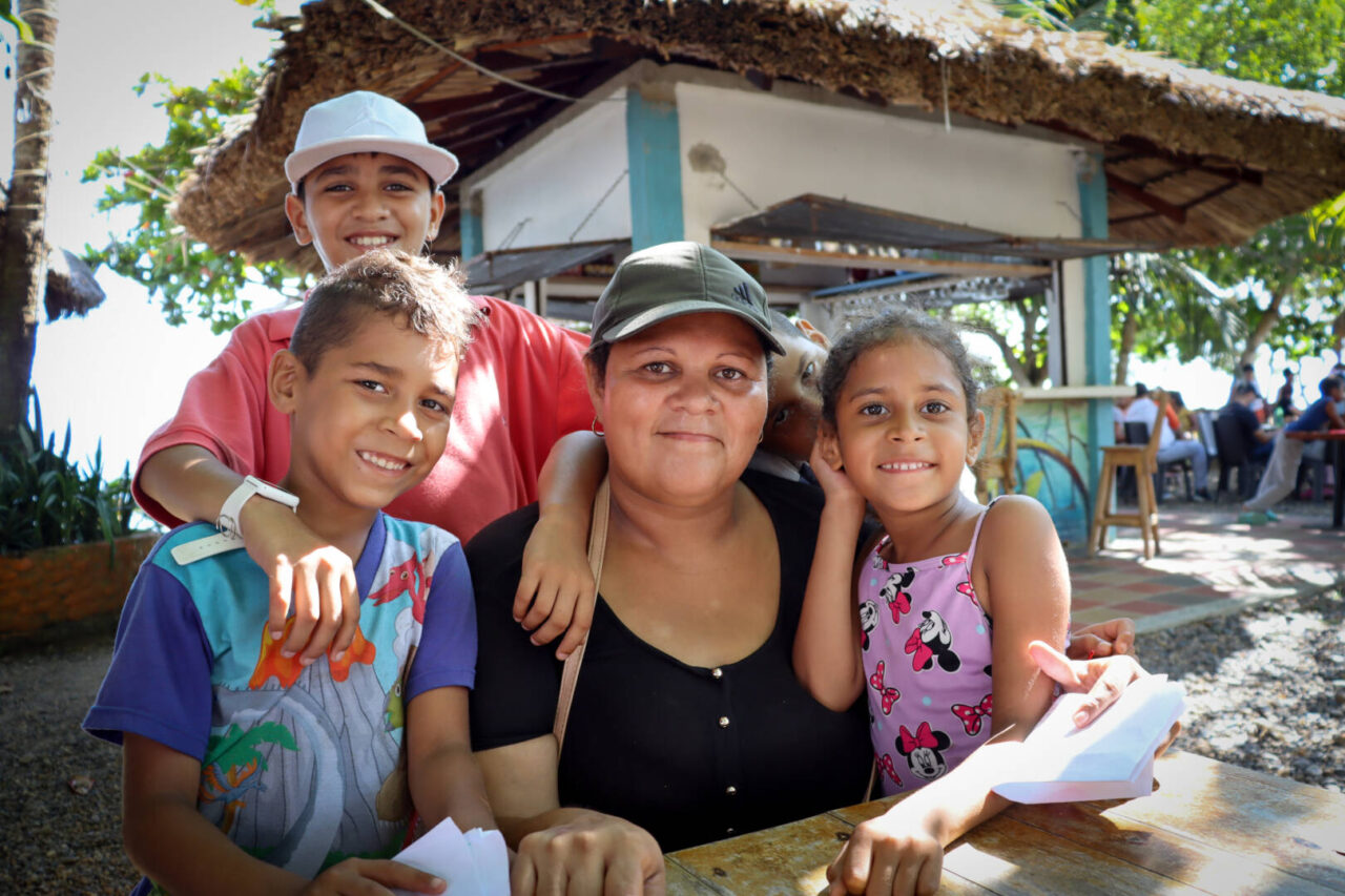 A woman wearing a baseball cap sits at a table in the shade, surrounded by her three children. They are all smiling.