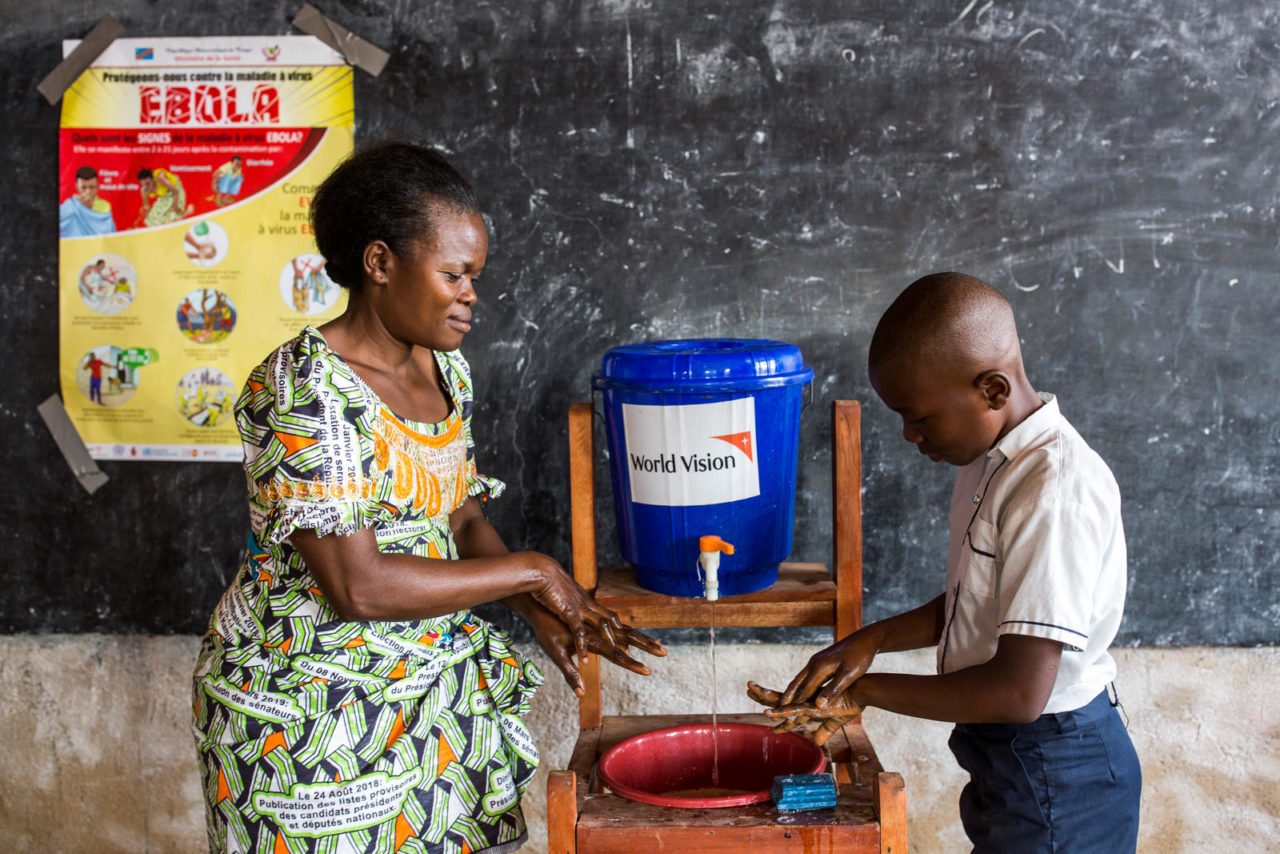 A teacher demonstrates handwashing to a student, using water from a blue World Vision bucket. A poster with information about Ebola is behind them.