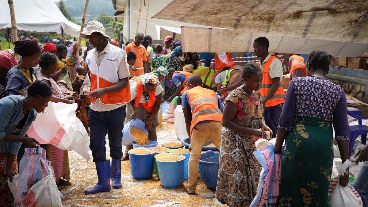 Men in orange vests help people carrying bags and blue buckets during a crowded food distribution.