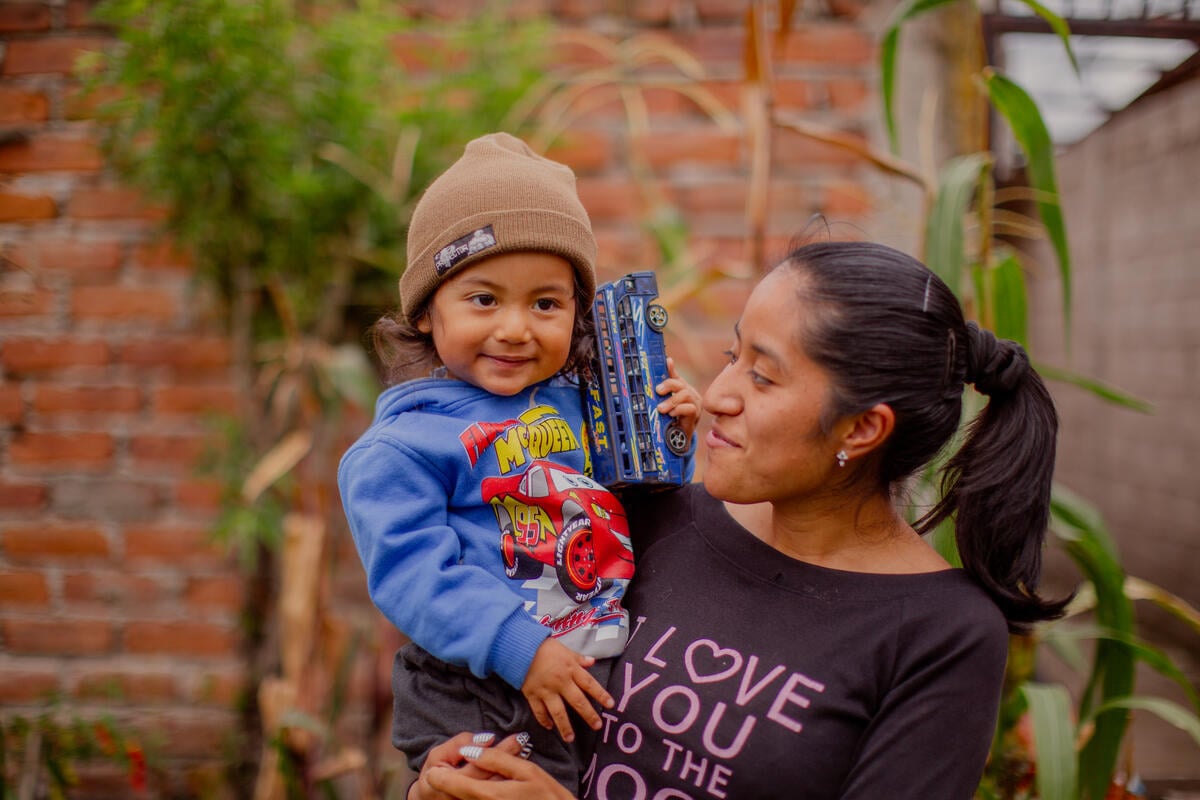 A smiling young boy holds a toy car while being embraced by his mother outdoors.