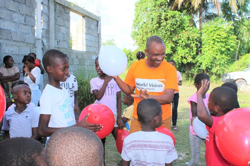 Surrounded by children, an aid worker in an orange World Vision T-shirt sports a warm smile as he grasps a white balloon.