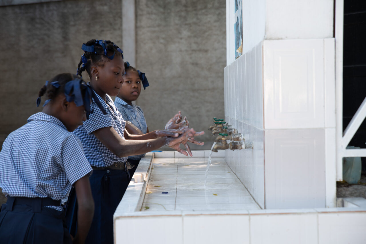 Three Haitian schoolgirls wash their hands at an outdoor handwashing station. Wearing ribbons in their hair, they sport blue gingham shirts.
