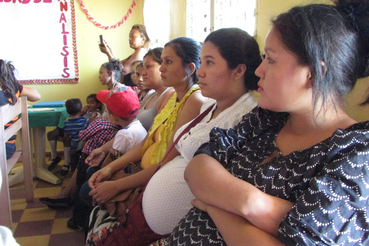  profile view of pregnant women sitting in a row, gazing straight ahead in a classroom with children. 