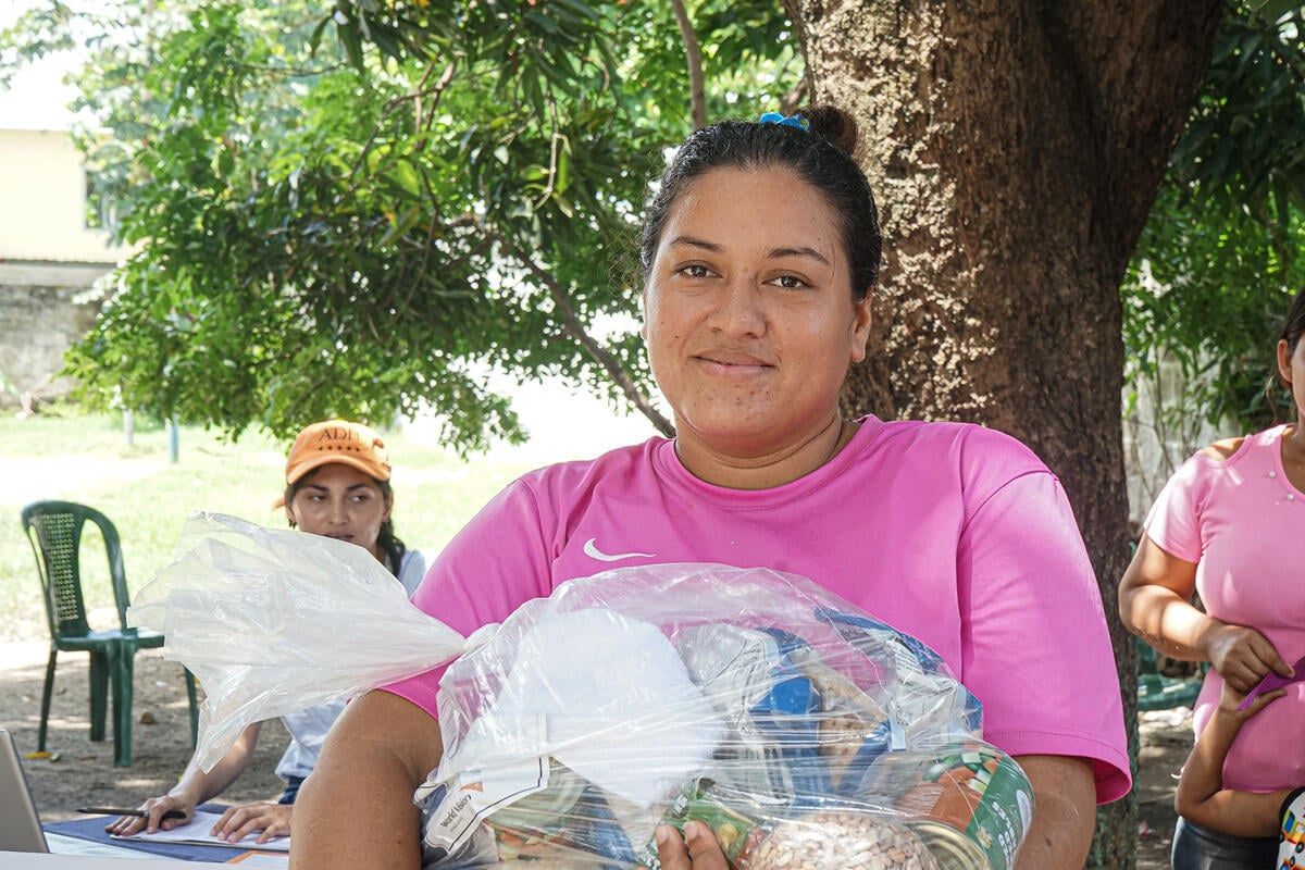 A woman in a pink shirt holds a large plastic bag of food supplies while sitting outdoors under a tree.