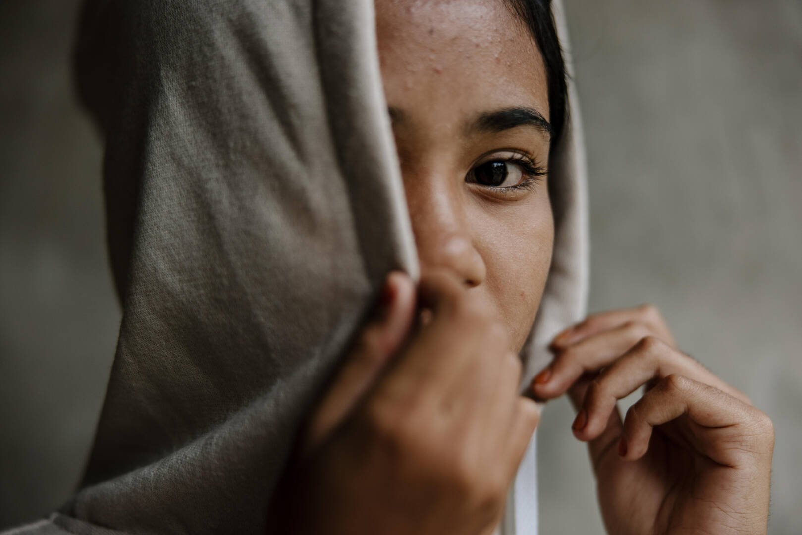 A teenaged girl covers part of her face with her hood and hand.