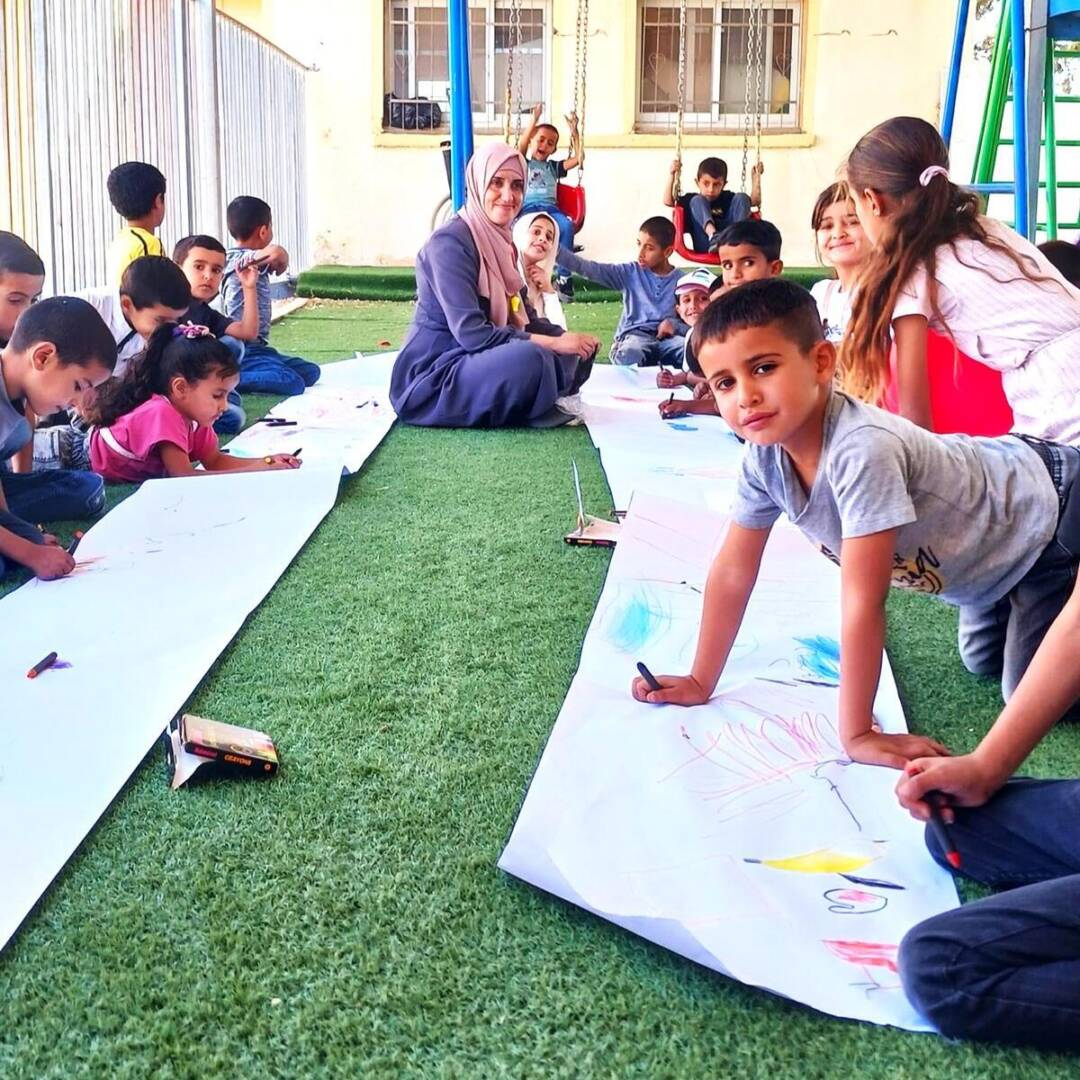 A boy looks into the camera, pausing from drawing on a roll of paper spread across the turf. Rows of children sit on the ground.