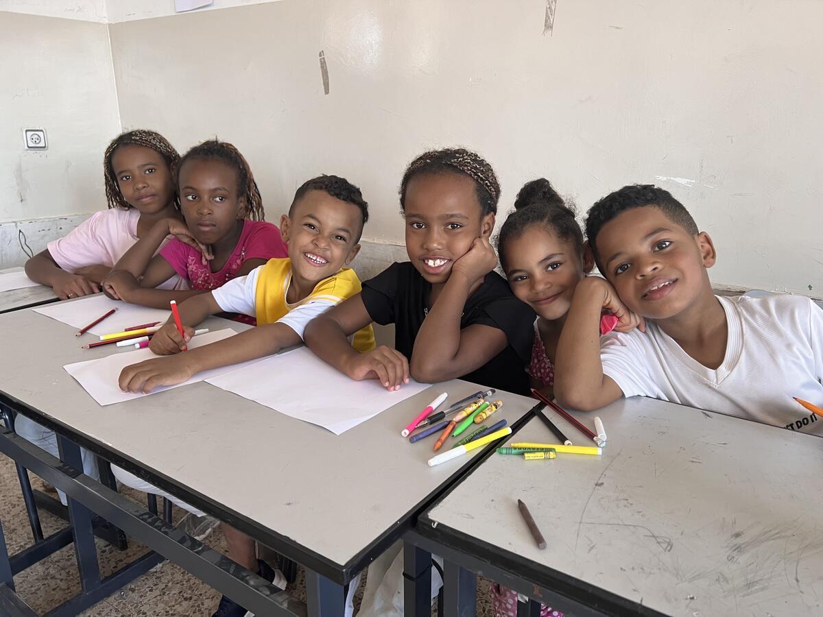 A row of children sitting at desks smile at the camera. Markers are lined on the desks, and some have sheets of paper on them.