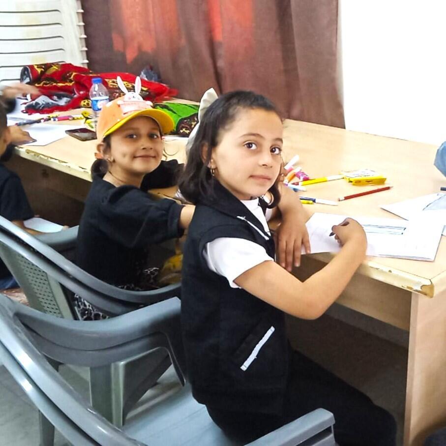 Two girls, one wearing a yellow baseball cap, glance up at the camera with smiles, taking a moment away from drawing.