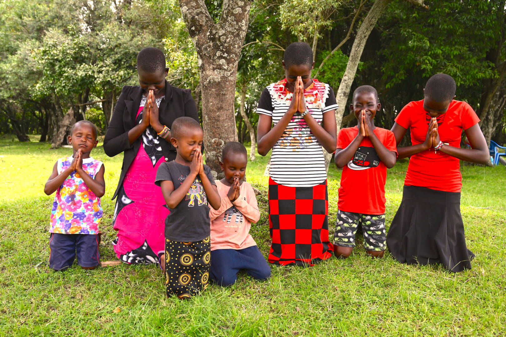 Seven children, with eyes closed and hands held in prayer, kneel on green grass near trees in Kenya.