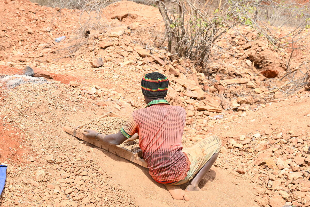 A child wearing a black knit cap sits with their back turned on dirt, breaking rocks in a Kenyan minefield.