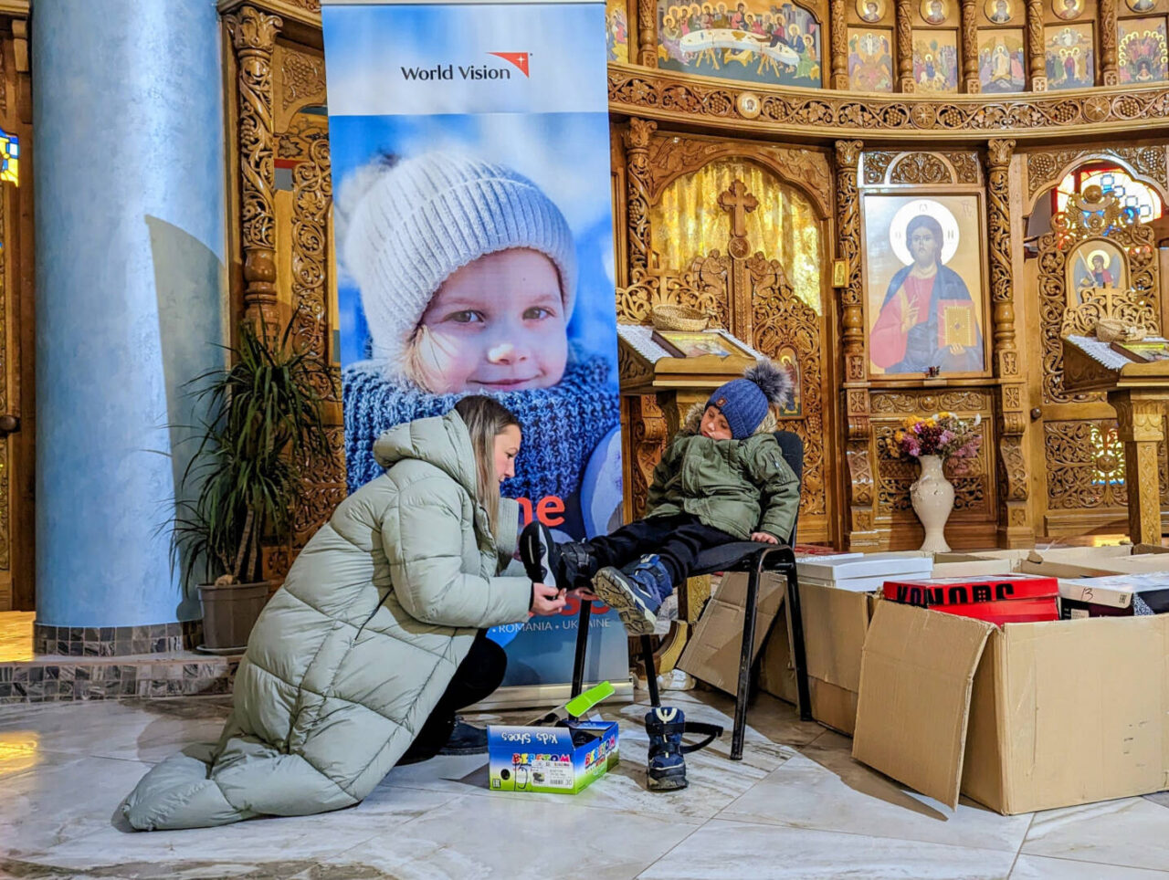 A little boy sits on a stool surrounded by cardboard boxes inside a golden-walled church. He is bundled up in a thick winter coat and hat. His mother crouches in front of him as she slips winter boots onto his outstretched feet.