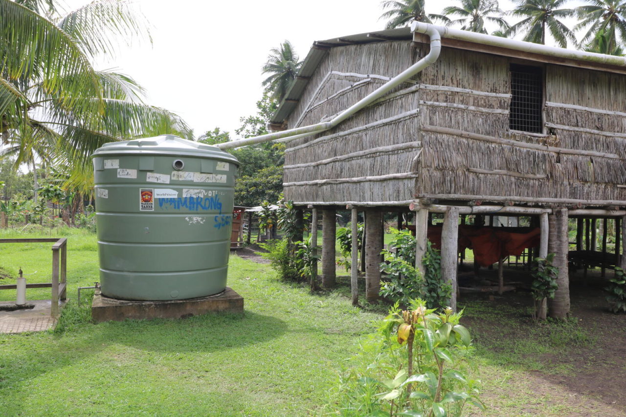 A pipe runs from a traditional home built on stilts over lush green land to a large water tank in a tropical setting. 