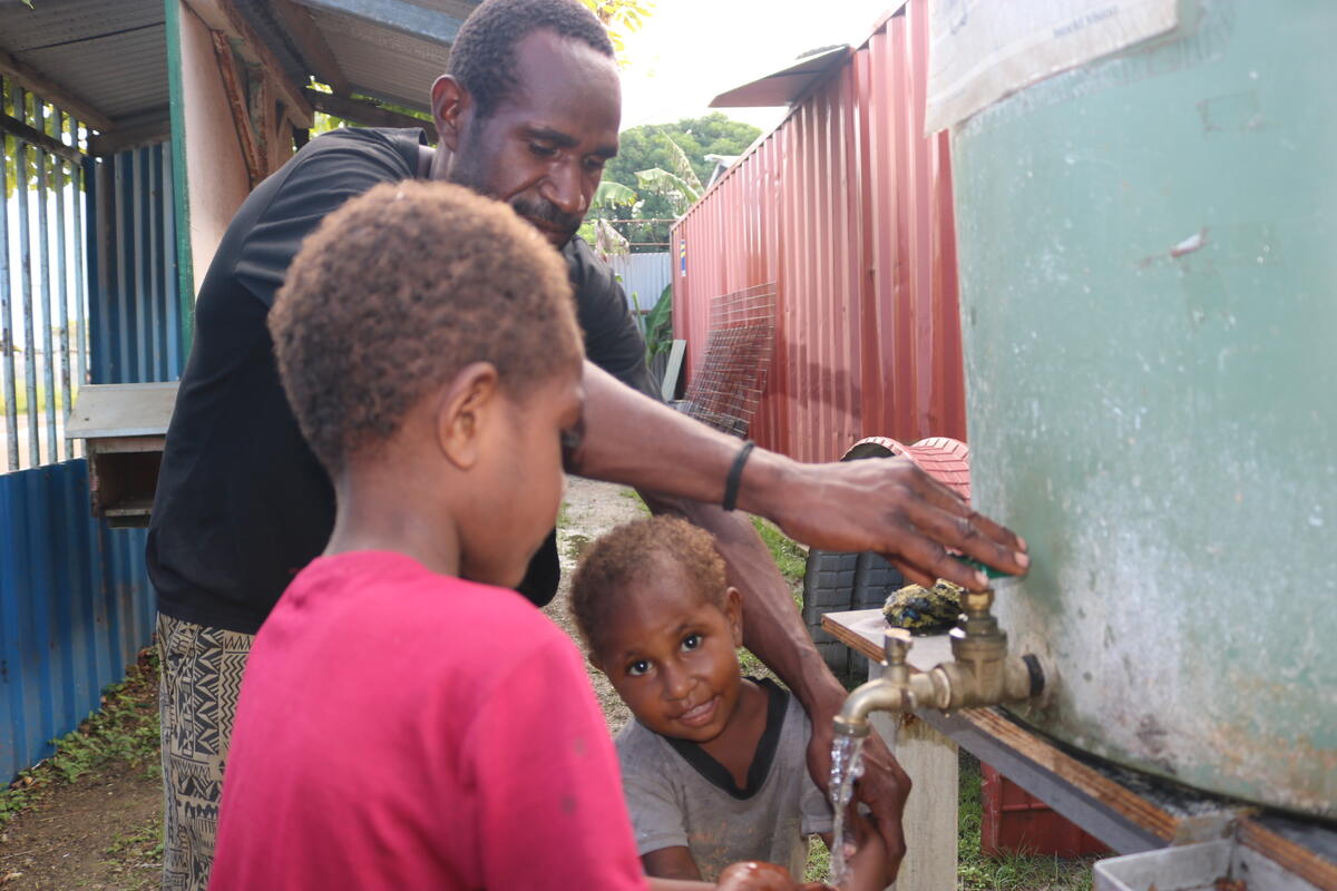 A girl smiles by a water tank as a man behind her places a hand on the knob and the other on her shoulder. A boy joins them.