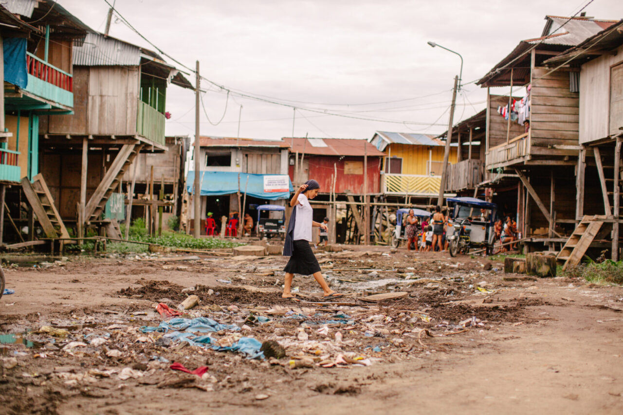 A young woman stands outdoors in a neighborhood. Behind her are houses raised on stilts. The dirt ground is scattered with debris.