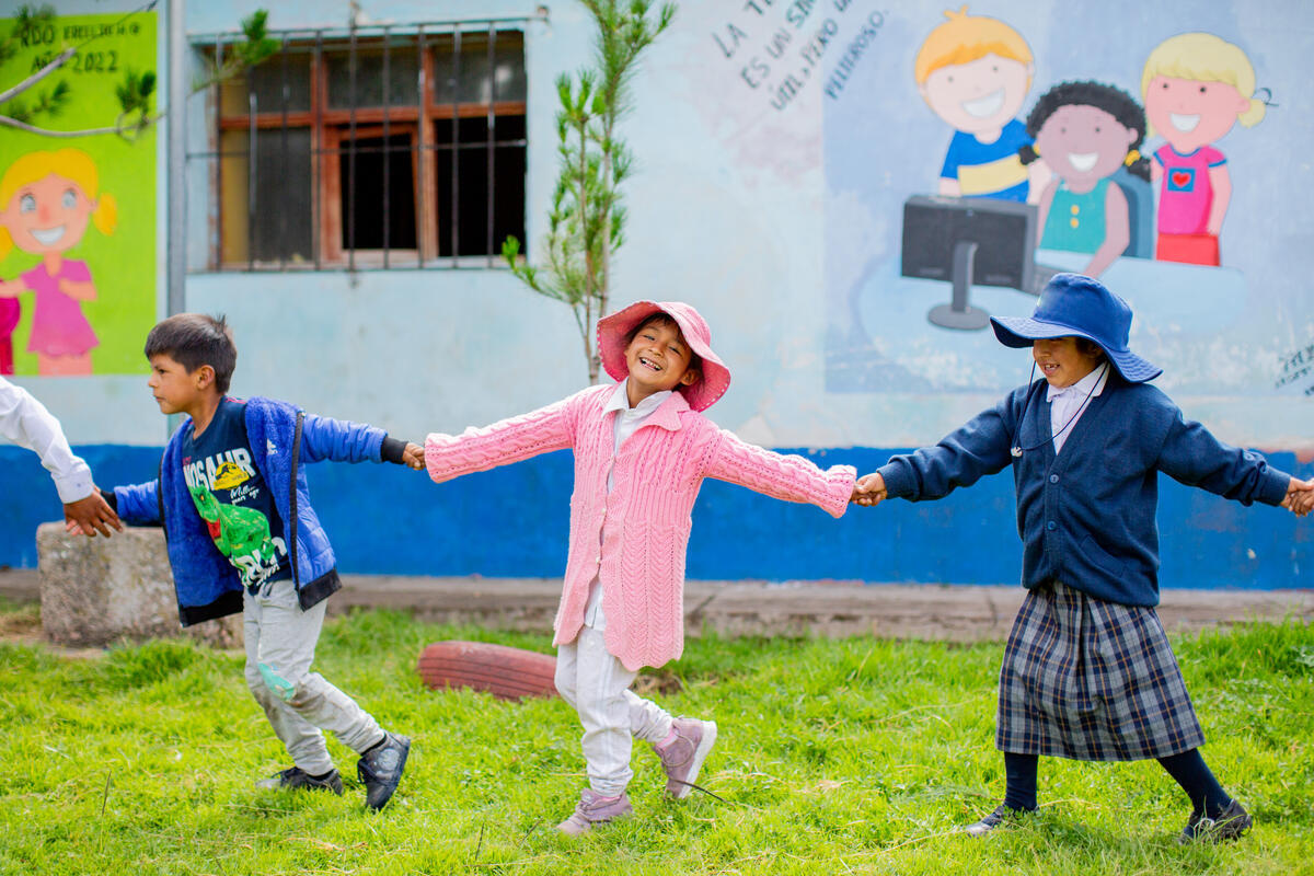 Three children, including a smiling girl in a pink hat and sweater, hold hands during a playground game.