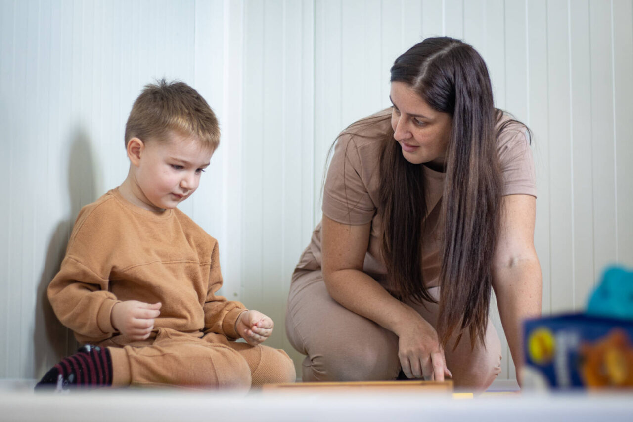 A young boy in a tan sweatsuit and a woman with long brown hair play with toys.