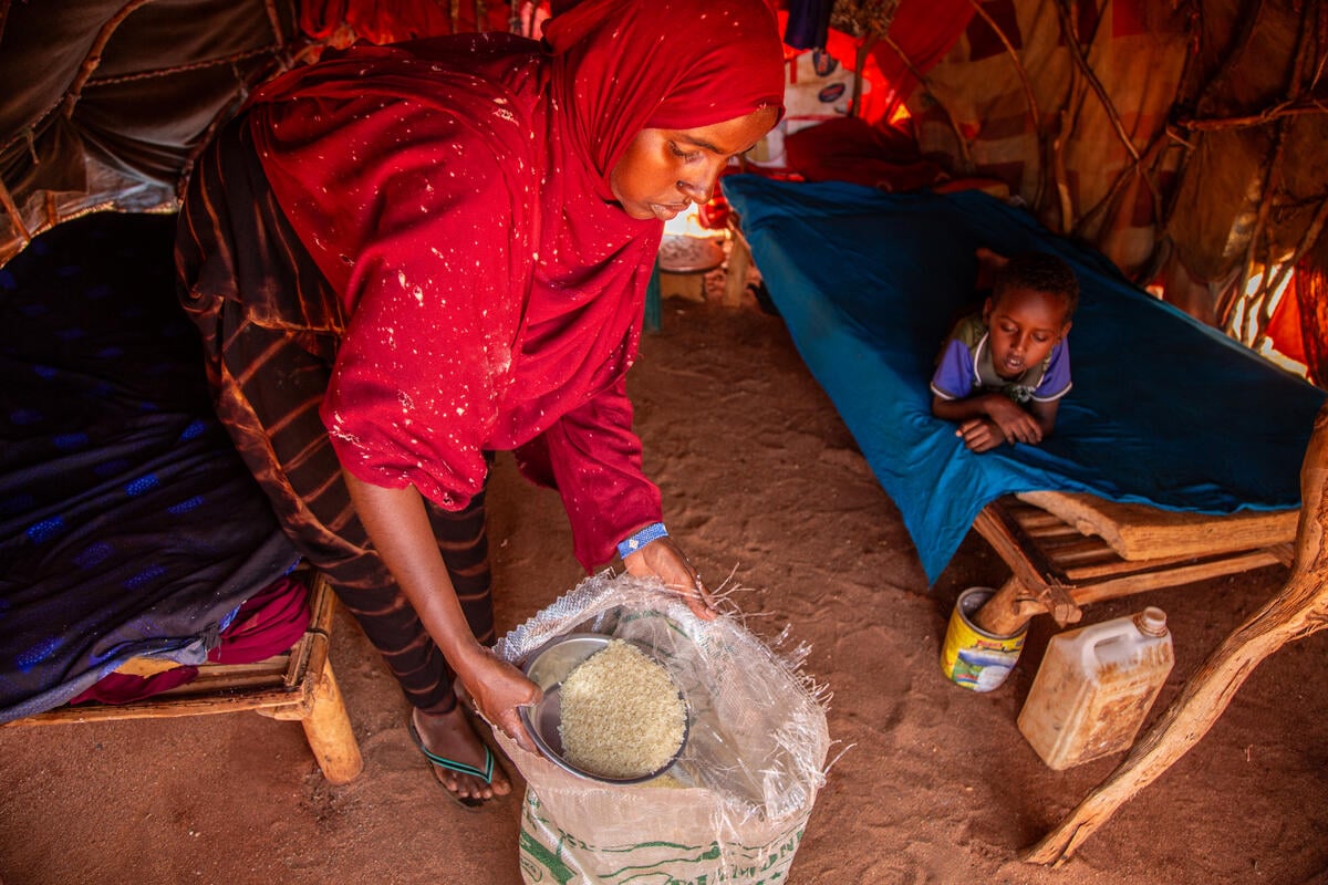 A woman wearing a red veil bends inside a tent, scooping a bowl of rice from a bag. Nearby, a boy lies on his stomach on a wooden bed.
