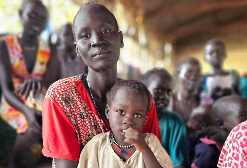 A woman holds a young child on her lap, both looking at the camera, with a blurred crowd in the background.