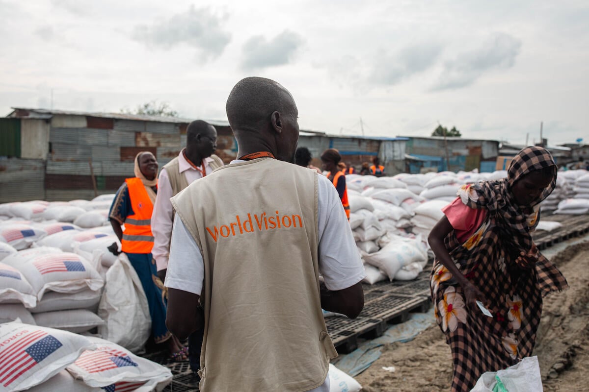 A man in a World Vision vest has his back to the camera. A woman and aid workers in orange stand among large food sacks with the U.S. flag.