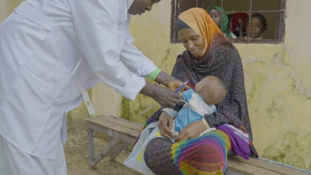 A health worker holds paper tape and a pen while reaching for a baby in a mother&rsquo;s lap.