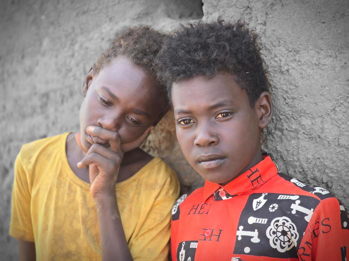 Two children side by side against a rough wall, one looking down with their hand to their face, the other gazing forward.