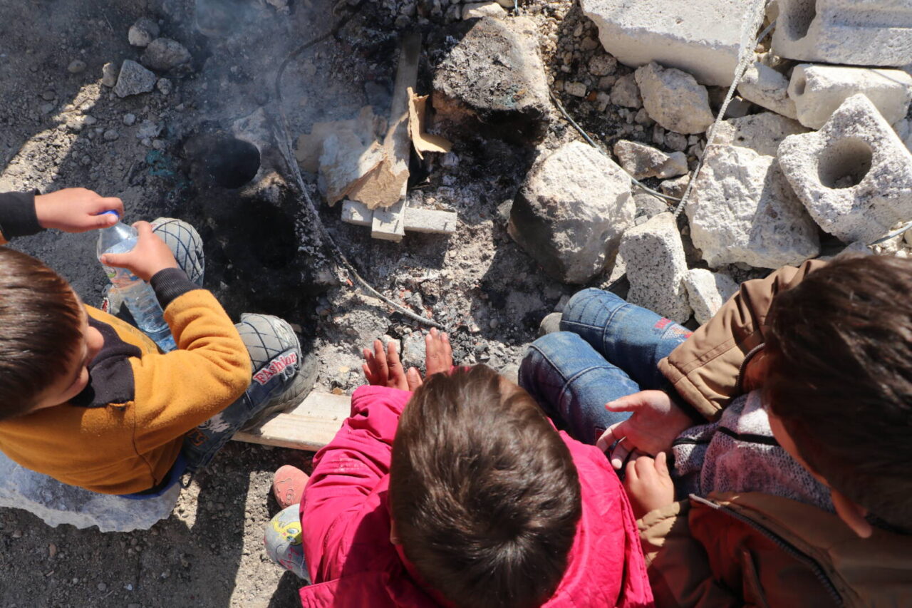 A bird&rsquo;s-eye view of three children in Syria sitting on rubble in front of a small smoldering fire.