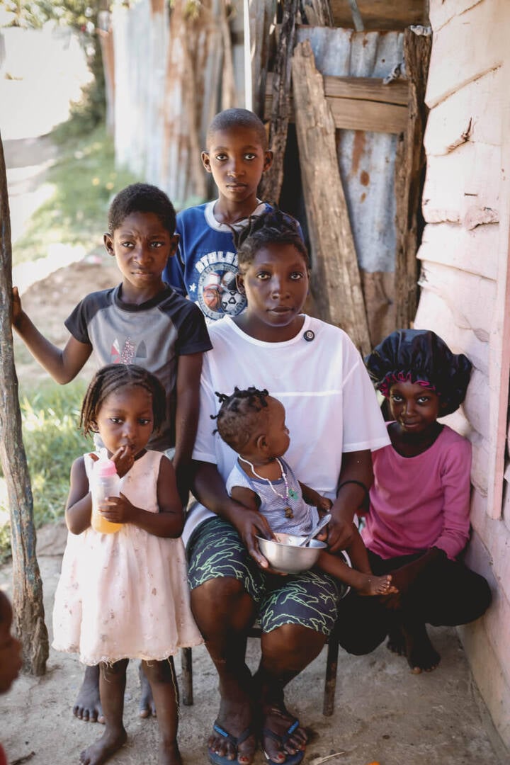 A woman in a white blouse gazes into the camera, surrounded by five young children. Four of the children look directly at the camera as they sit outside near a wooden structure. 