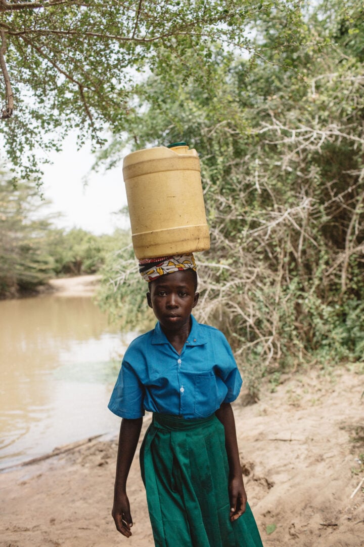 A girl carries a bucket of water on her head as she walks away from a dirty pond.