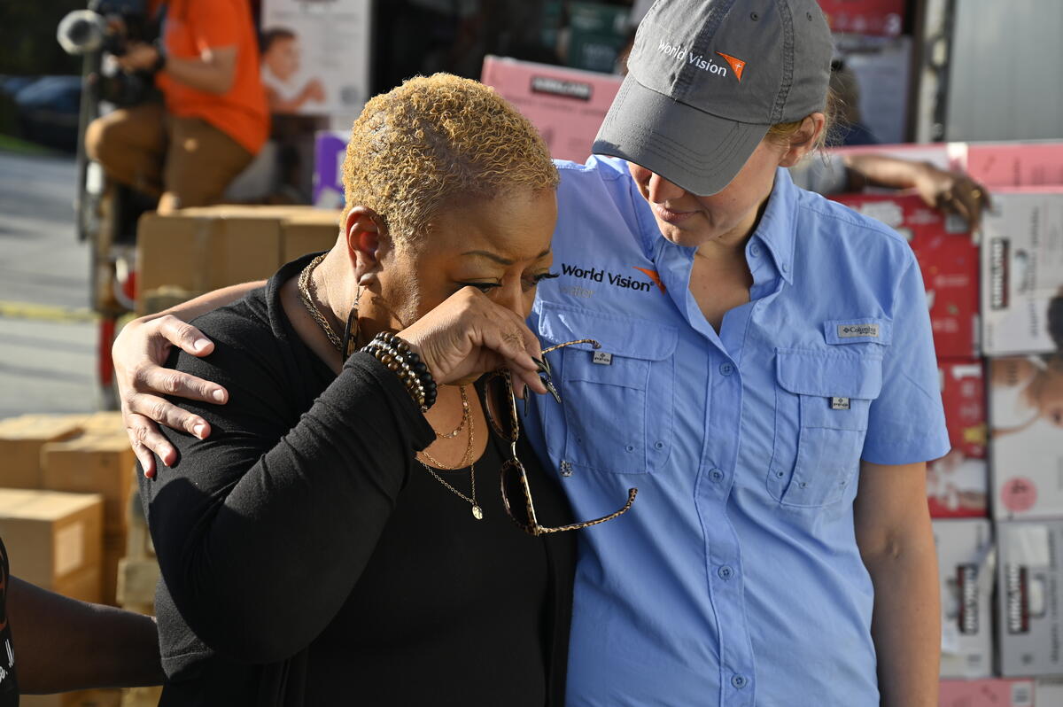 A woman stands with her arm around another woman who wipes her eyes. Boxes are stacked behind them.