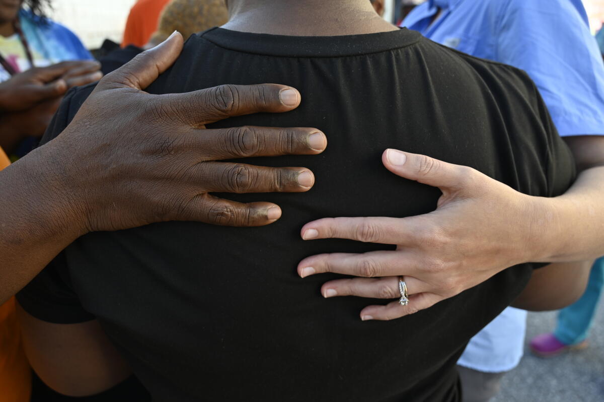 Hands rest on the back of a woman wearing a black T-shirt in a gesture of support during prayer. 