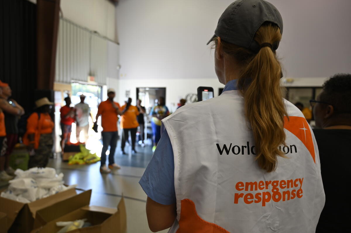 A woman stands with her back to the camera, facing a group gathered inside a gym. Boxes and piles of relief supplies are on the gym floor.